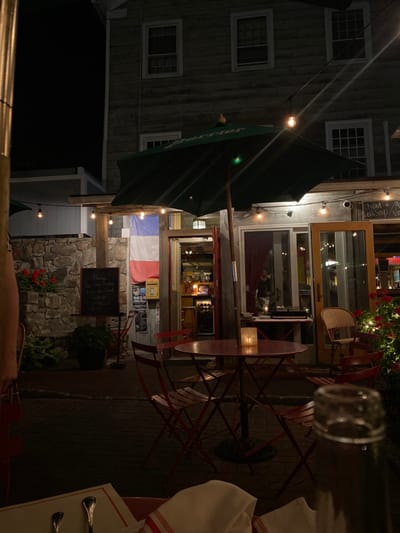 The image depicts an outdoor patio of a restaurant at night, specifically identified as Luc's Cafe located at 3 Big Shop Lane in Ridgefield, Connecticut, United States. The scene is illuminated by artificial light sources.
A multi-story building, likely the restaurant structure, occupies the background, featuring light-colored horizontal wooden siding and numerous rectangular windows. A prominent French flag is vertically displayed near the main entrance. The entrance consists of a glass-paned double door on the right and an open doorway on the left, leading into illuminated interior spaces where a blurred figure, possibly a male, is partially visible through the glass door.
The patio area is furnished with multiple red metal bistro-style tables and matching folding chairs, arranged on a dark paved surface. A large, dark green patio umbrella with "Perrier" branding is centered over one of the tables, beneath which a white candle in a holder provides illumination. String lights with exposed bulbs are strung across the patio, providing overhead illumination.
To the left, a stone wall supports a planter containing red flowers and green foliage. A black chalkboard sign with white text, partially obscured but reading "Please see the Hostess When you enter," stands near the entrance. A yellow, mailbox-like object is affixed to the wall beside the French flag. To the right of the main entrance, a white board with handwritten text (e.g., "Chilled A...", "Corn Soup...") is visible on the building wall, and a security camera is mounted above the right door frame. Additional red flowers and string lights are present on the right side of the patio.
In the immediate foreground, a white napkin with red stripes and silverware (a fork and spoon) are visible on a table, implying the perspective of a diner. The top of a clear glass bottle is partially visible on the far right of the foreground table. The overall environment is dark, typical of nighttime, with artificial lighting creating an intimate atmosphere.