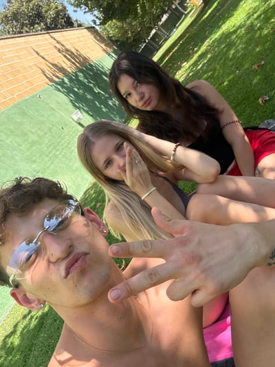 In the city of Villa Del Prado, Spain, three young adults are captured in a close-up, low-angle selfie outdoors. The scene takes place on a grassy area adjacent to a green artificial turf surface, likely a sports court or recreational area, with a brick-patterned wall visible in the background casting dappled shadows from overhead trees.
In the foreground, a young man with curly brown hair and sunglasses is prominently featured, looking towards the camera. He is shirtless and wearing a thin gold chain. His right hand is raised, making a peace sign gesture, and his thumb is touching his index finger. He has a nose piercing and earrings.
Behind him and to his right, a young woman with long blonde hair is partially visible. She is wearing a dark top and has her hand raised, covering her mouth and nose, with rings on her fingers. She has blonde bracelets on her wrist.
Further back and to the right, a young woman with long dark hair is also visible, wearing a black top and red shorts. Her arm is resting on her leg, and she has a beaded bracelet on her wrist.
The lighting suggests it is daytime, with sunlight illuminating the subjects and casting shadows. The overall atmosphere appears relaxed and casual, indicative of an outdoor social gathering.