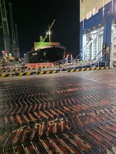 The image shows a nighttime scene at a port, likely in Ravenna, Italy, with a large cargo ship named "GULLUK" docked. In the foreground, a textured, dark surface with red and grey markings covers the ground, possibly a ramp or loading area. To the right, a large vessel with blue and white sections and open rampways is partially visible. In the mid-ground, the bow of the "GULLUK" ship is illuminated, revealing its name and hull details. Tall cranes and port infrastructure are visible in the background, silhouetted against the dark night sky. The scene suggests ongoing port operations or preparations for loading/unloading. There are no people clearly visible in the image.