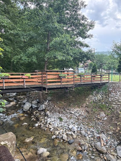 This image captures a tranquil outdoor scene in Polena, Bulgaria, featuring a narrow stream or small river flowing through a natural yet somewhat developed area. The perspective is slightly elevated, looking down towards the water and across to the opposite bank.
In the foreground and to the right, a rocky stream bed is visible, with clear water gently flowing over various sizes of grey and brown stones and pebbles. Some small ripples and turbulent sections indicate current. A black pipe runs parallel to the stream along the bank, and a concrete structure is partially visible in the bottom right corner. The stream bank on the right is composed of bare earth and loose rocks, with sparse patches of green vegetation.
To the left, a dense canopy of green trees with abundant foliage dominates the scene, suggesting a lush, wooded environment. A prominent wooden structure, resembling a rustic fence or railing, runs horizontally across the middle of the frame, parallel to the stream. This structure appears to be part of a footbridge or an elevated walkway. It is constructed from brown wooden planks and slats, supported by metal elements and wires. Several small terracotta-colored plant pots, holding green plants, are hanging from the wooden railing, adding a touch of domesticity to the natural setting.
In the background, beyond the wooden structure and through the trees, glimpses of distant houses or buildings can be seen, hinting at a village or settlement. A light-colored vehicle, possibly a white truck or van, is partially visible parked in the distance. Further back, a stone wall and a green wire fence are also discernible. The sky above is overcast with soft grey clouds, indicating a cloudy or partly cloudy daytime setting. No people are visible, and no discernible text appears in the image. The overall atmosphere is peaceful and serene, blending natural elements with subtle signs of human presence.