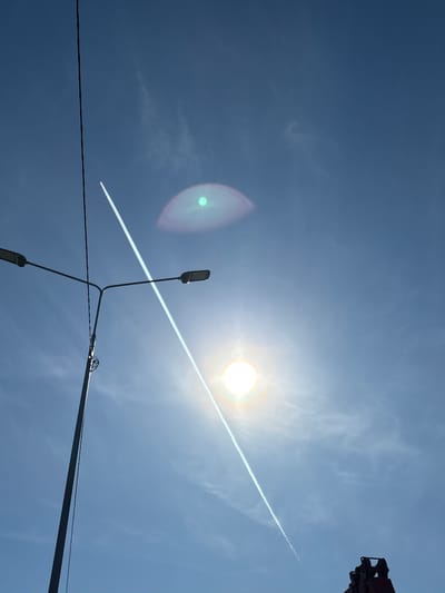 The image shows a bright, sunny day in Verkh-Sosnova, Russia. The sky is a clear blue, with a vapor trail from a jet airplane crossing the frame diagonally. The sun is visible, causing a lens flare effect. In the foreground is a lamppost with two lamps extending at different angles. Electrical wires are also present, crossing the lower part of the image. The upper right corner shows part of some metallic machinery. There are no people visible.