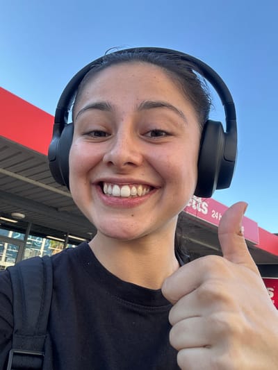 A young woman is taking a selfie outdoors. She is smiling broadly and giving a thumbs-up. She is wearing black over-ear headphones and a black t-shirt with a strap across her shoulder, possibly from a bag. The background includes a red awning with white writing, "24h". The sky is blue. Behind her, one can see reflections of buildings in what appears to be windows or glass doors. Considering the presence of the "24h" sign and the window reflections, this image was likely captured at a gas station convenience store or a similar retail establishment. While Applecross in Australia can't be definitively confirmed solely from this photo, the details suggest a roadside setting potentially along Canning Highway in Applecross, where some 24-hour stores are located.
