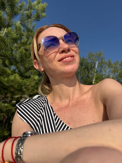 A woman with blonde hair pulled back in a ponytail, wearing blue-tinted sunglasses and a black and white striped top, smiles as she looks up at the sky. She has on a hoop earring in her left ear and a watch on her left wrist, along with a red string bracelet. The background consists of green pine trees, other foliage, and a clear blue sky, suggesting an outdoor setting, possibly in Shuiskaya Chupa, Russia.