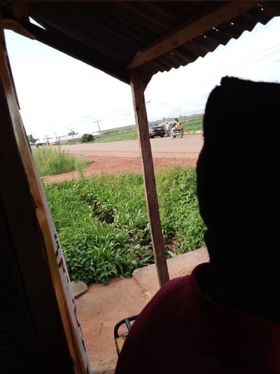 The image captures a scene viewed from under a rustic, corrugated metal roof supported by wooden beams. In the foreground, the silhouette of a person wearing a dark red shirt is visible, obscuring a large portion of the right side of the frame. The person's head and shoulders are a solid black shape against the brighter background.

Beyond the immediate foreground, a dirt road cuts through a landscape dotted with lush green vegetation. In the middle ground, a person is riding a motorcycle, and next to them, a car is parked. Two other individuals stand near the car. The road leads towards a cluster of buildings in the distance, with several utility poles visible, suggesting a semi-rural or village setting. The fields surrounding the road appear to be cultivated.

The sky is overcast, indicating a cloudy or possibly rainy day. The overall lighting suggests it is daytime, though the exact time is difficult to determine. The presence of the road, buildings, and people on vehicles points to everyday activity in a Nigerian village. No visible text is present in the image. The scene conveys a sense of ordinary life unfolding in a tropical or subtropical environment.