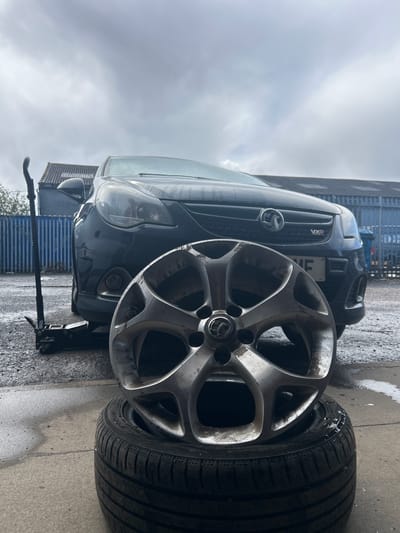 The image depicts an outdoor vehicle maintenance scene, likely within an industrial or commercial estate in Rutherglen, United Kingdom. In the foreground, a dirty, silver-colored, multi-spoke alloy wheel, bearing a Vauxhall emblem in its center cap, is stacked on top of a black Yokohama tire. Both the wheel and tire appear wet. In the midground, a dark-colored Vauxhall Corsa VXR, identified by its grille badge and UK registration plate partially visible as "ME", is elevated on its front driver's side by a black hydraulic floor jack. The car's windshield is wet. The ground surface is wet concrete or asphalt. The background features a tall blue corrugated metal fence, behind which a building with a grey corrugated roof is visible. Another building with a similar roofline is partially visible to the far left. The sky is overcast and grey. No individuals are present in the scene.