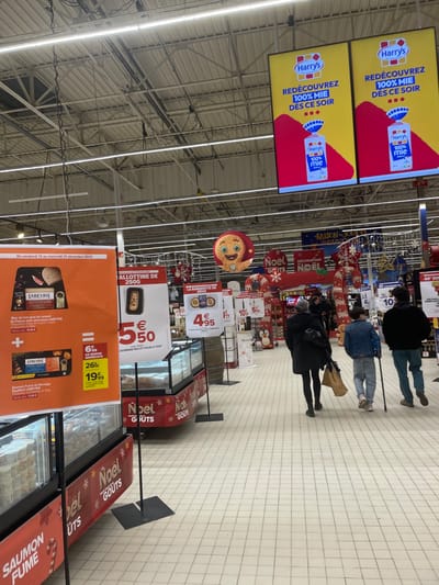 The image depicts a grocery store aisle with advertisements and Christmas decorations. Above the aisle, there are two screens displaying a Harry's bread advertisement with the text "REDÉCOUVREZ 100% MIE DES CE SOIR". Below, Christmas-themed decorations adorn the aisle. Product displays feature promotions for "LABEYRIE" including foie gras, and "SAUMON FUME" with prices shown. A sign promotes a "LA BARQUETTE DE" with a price of €4.95. Shoppers are visible further down the aisle.