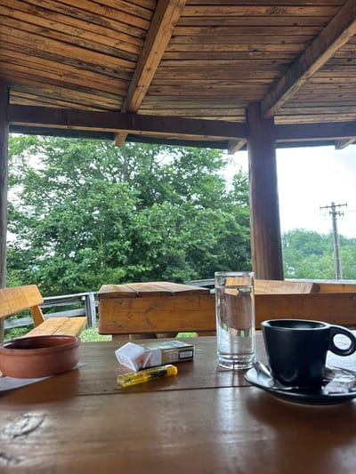 This image shows a close-up view of a wooden table with several items on it, set against a backdrop of lush green trees and a bright sky. The table appears to be located outdoors, possibly on a porch or patio, as indicated by the wooden structure and the natural surroundings.
On the table, there is a black ceramic coffee cup and saucer, a clear glass filled with water, a packet of cigarettes, and a small yellow object that resembles a lighter or pen. A brown ceramic bowl is also visible. The wooden structure in the foreground, with its rough-hewn planks, suggests a rustic or natural setting.
The time of day appears to be daytime, with ample natural light. The sky is overcast, suggesting possibly cloudy weather. The presence of trees indicates a location with abundant vegetation, and the overall scene evokes a sense of relaxation and casual enjoyment, perhaps a moment of pause during a visit to Mekića Kula, Montenegro. There is no visible text in the image.
