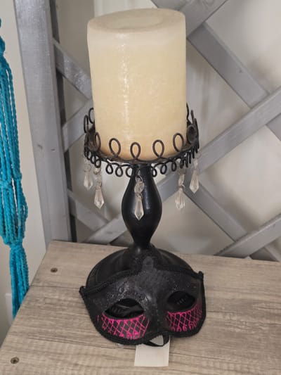 A still life image features a black, ornate candle holder with a tall, cream-colored pillar candle. Dangling from the candle holder are several clear, faceted crystals. At the base of the candle holder rests a black masquerade mask, adorned with pink diamond-shaped detailing and glitter. To the left, a portion of a bright blue, macrame-style decoration hangs, its tassels visible. The objects are placed on a light-colored, wooden surface, possibly a table or shelf. In the background, a grey, slatted wooden structure is visible, with hints of a white wall behind it. The lighting is soft and diffused. The image appears to be taken indoors.