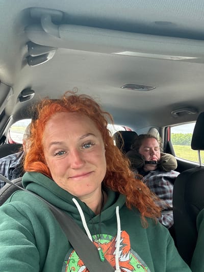 In the foreground of the image, a woman with vibrant red, curly hair is looking directly at the camera, smiling faintly. She is wearing a dark green hooded sweatshirt with a white drawstring and a colorful graphic design on the front. A seatbelt is visible across her chest. In the background, another person is sleeping in the back seat of a car, propped up by a travel pillow and appearing to be wearing a plaid shirt. The interior of the car is visible, with the ceiling and grab handles present. Outside the car window, a green, leafy landscape is visible under a cloudy sky. The location is stated to be Alnashi, Russia.