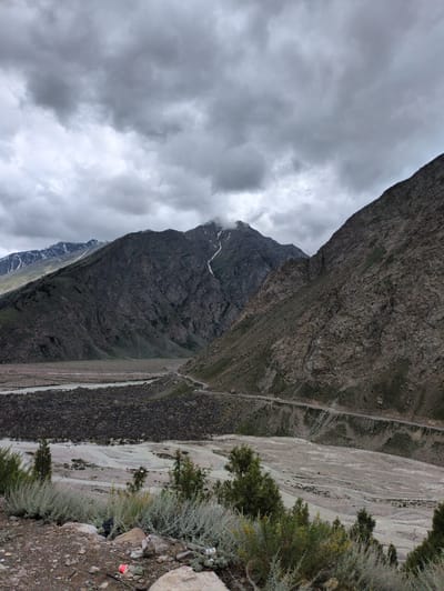 The image depicts a dramatic mountain landscape under a cloudy sky. In the foreground, there is a rocky, uneven terrain with sparse, low-lying vegetation and some scattered litter, including a red plastic cup and black plastic bags. A dusty road, possibly a mountain pass, winds its way along the side of a steep, rocky mountain face that dominates the right side of the frame.
Further back, a wide, dry riverbed or valley stretches across the middle ground. It is filled with pale, sand-colored sediment and scattered rocks, with a faint, meandering stream visible in parts. On the left side of the valley, another mountain range rises, its slopes covered in dark, rugged rock and patches of green vegetation. The peaks of these mountains are partially obscured by clouds, and snow or ice can be seen on the higher elevations. The overall atmosphere is one of a remote, rugged, and natural environment.
The location is identified as Darcha, India, a high-altitude settlement in the Lahaul and Spiti district of Himachal Pradesh. Darcha is known for its stunning mountain scenery and is a popular starting point for trekking routes in the region, including those leading to the Zanskar valley. The scene captured is consistent with the topography and environment of such a location.
