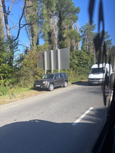 The image captures a roadside scene in Ianeti, Georgia, under a clear blue sky. A dark gray Jeep Patriot SUV, displaying license plate HH-644-GM, is parked on the right shoulder of an asphalt road, with its left wheels on the pavement and its right wheels on the grassy verge. Behind the Jeep, partially obscured by green and brown foliage, stands a large, multi-panel structure constructed from corrugated silver metal. Further ahead on the road, a white Mercedes-Benz Sprinter-style van, with license plate NJ-981-LM, is visible. The asphalt road features a solid white line marking its edge. Tall trees with slender trunks and varying densities of foliage line the right side of the road, forming a dense background. Shadows are cast across the road surface from a light source positioned to the left. No individuals are visible in the frame. The image appears to be taken from inside a vehicle, with parts of the right foreground obscured by a window frame or reflection.
