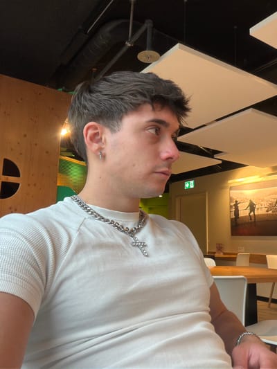 A profile view of a Caucasian male wearing a white shirt and a silver chain with a cross pendant, likely in a cafe or restaurant. He is wearing a silver bracelet and a small earring. The background shows a play area with green netting and wooden structures, and geometric ceiling panels. In the distance is a framed picture on the wall. The location is likely a restaurant or cafe in Lucerne, Switzerland, possibly with an indoor play area for children.