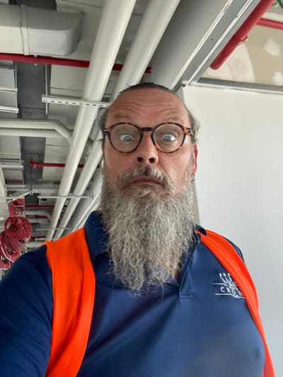 A close-up, tilted shot shows a man with a full, gray beard and round, tortoiseshell glasses. He is wearing a navy blue shirt with an orange safety vest layered over it. A logo is visible on the chest of the shirt, appearing to be embroidered in white thread and reading "CROWN". The man's expression is neutral. The background consists of industrial piping, mostly white, with some red accents and red spools of wire visible towards the bottom right of the frame. The setting appears to be an indoor industrial or construction environment. The lighting suggests it is daytime, and there are no clear indicators of the weather. The orientation of the image is rotated 90 degrees counter-clockwise.