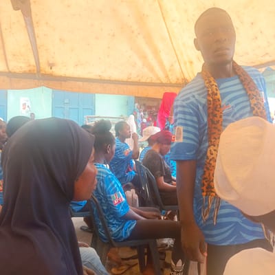 A group of people are gathered under a large, weathered yellow-tan tent. Most individuals wear blue shirts with a logo visible that says "APC." Several people sit on black plastic chairs. The background shows a blue building with closed doors and a poster on the wall. A man stands in the foreground wearing a blue shirt with a leopard-print scarf. The location is Gudi, Nigeria, based on the shirt logos that contain "Nasarawa North Senatorial District."