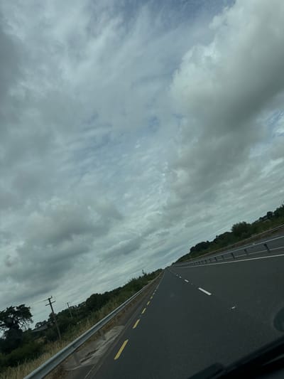 The image shows a view from inside a vehicle traveling on a highway. The road is dark asphalt with white lane markings and yellow solid lines on the side. There is a metal guardrail along the left side of the road, separating it from a grassy embankment with some trees and vegetation. In the distance, more trees and rolling hills are visible under a cloudy sky. Two utility poles are visible on the left side of the road. The sky is filled with various shades of gray clouds, indicating an overcast day. The perspective suggests the vehicle is moving forward, away from the viewer. The location is likely a rural area within the New Ross Municipal District of Ireland, given the typical road infrastructure and landscape. There are no people or other vehicles visible in the frame.