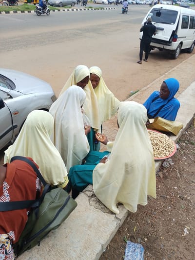 The image depicts an outdoor scene with several people, likely in Nigeria, near a road. In the foreground, a group of individuals, predominantly women and girls wearing hijabs, are gathered. One person in a blue hijab is seated next to a large platter filled with peanuts. Another person, also in a hijab, is holding a handful of peanuts, possibly in the process of selling or distributing them. Several other people in light-colored hijabs are looking at the peanuts or engaged in conversation.
To the left of the group, part of a silver car is visible. Behind the car, the road stretches out, with various vehicles, including motorcycles and a white van, present. A man in a black shirt and trousers is standing near the open back of the white van, appearing to be on his phone.
The setting appears to be a marketplace or roadside area, indicated by the presence of goods for sale (peanuts) and the proximity to a road with traffic. The ground is a mix of dirt and gravel. The lighting suggests it is daytime, possibly overcast or late afternoon, as there are no harsh shadows. The overall mood seems to be one of commerce and everyday activity. No visible text is clearly legible in the image.