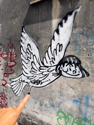 A piece of street art depicting a white bird-like character with black outlines is spray-painted on a weathered concrete wall. The character features an animated face with large eyes and a hat-like structure on its head. It has a wing spread out to the left. To the lower right of the graffiti, a finger with a dark-polished nail points toward the artwork. There is another piece of graffiti with a maroon outline below and to the left of the bird. The concrete wall has a rough texture and shows signs of aging, with various discolorations, spots, and other markings. There is green graffiti visible to the upper right of the bird. The scene suggests an outdoor environment, likely a part of the city of George Town, Malaysia, known for its street art.