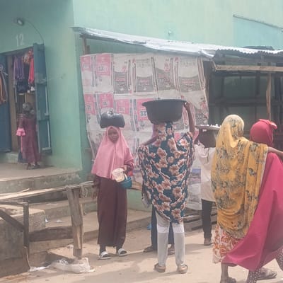In Gudi, Nigeria, a street scene shows women engaged in commerce. One woman with a pink headscarf carries a container and a bag on her head. Another woman in a floral print headscarf is carrying a bowl on her head. A woman wearing a yellow and red headscarf is walking by. Behind them, "Dangote" cement bags cover a structure, possibly a shop. A building painted in green bears the number "12," where clothing hangs. Litter is on the ground near the steps.