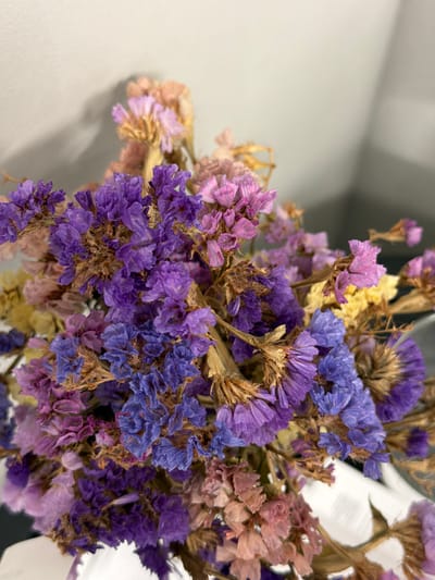 The image shows a close-up of a bouquet of dried flowers in varying shades of purple, pink, and yellow. The flowers are densely packed, with visible stems and filler materials. The background is a neutral grey surface on the left and a white object on the right. The image appears to be taken indoors under soft, diffused lighting. No information is available about the location in the city of Zagarancea, Moldova.