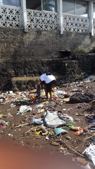 The image depicts a scene of pollution and cleanup efforts at a coastal area in Limbe, Cameroon, located at coordinates 4.005 latitude and 9.21 longitude. A person wearing a white shirt and black pants is seen bending over, picking up garbage from the sandy ground. The area is littered with a large amount of debris, including plastic bottles, bags, food wrappers, and other miscellaneous items. The person appears to be part of a cleanup initiative, as the image portrays their actions of gathering and removing trash from the ground. The background features a concrete structure with decorative patterned walls and a partly visible roof. The structure sits on a rocky cliff, with vegetation growing sparsely on the slope. The ground is covered with sand and debris, suggesting a beach or waterfront location.