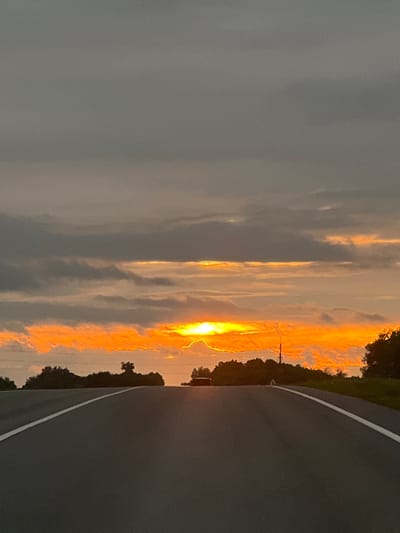 The image depicts a highway at dusk, with the sun setting in the distance. The sky is filled with dramatic clouds, illuminated by the vibrant orange and yellow hues of the sunset. The road, a dark asphalt surface, is marked with a white line that stretches into the horizon. Trees and a distant structure are visible on either side of the road, silhouetted against the colorful sky. A dark-colored vehicle is present on the road ahead, further down the highway. The overall scene conveys a sense of journey and the beauty of a fading day. While the image captures a general landscape, specific details about it being located in the city of Lermontovskii, Russia, are not visually present within the provided image.