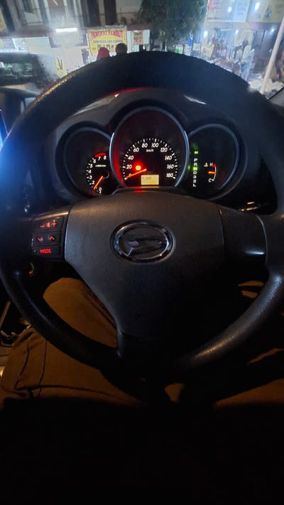 The image shows the interior of a car, focused on the steering wheel and dashboard. A digital odometer displays "174202". The gear selector shows "P R N D 3 2 L" with "N" illuminated in green, indicating the car is in neutral. The steering wheel has control buttons, including a "MODE" button. In the background, a sign reads "PANGKAS RAMBUT JHON PARIS JAYA PARDOSI", which translates to "Jhon Paris Jaya Pardosi Hair Salon". This sign is characteristic of businesses found in Batam, Indonesia. The overall lighting suggests it is nighttime.