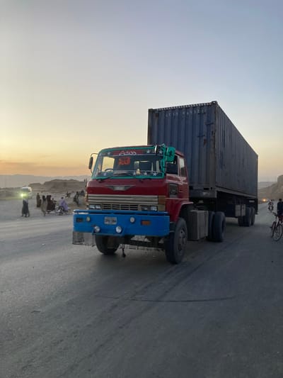 The image shows a truck carrying a shipping container on a road in Kuchlak, Pakistan. The truck has a red cab with blue accents and decorative elements. The container is dark-colored and stacked on the truck's trailer. There are people and vehicles, including motorcycles, in the background. The environment includes a road, mountains, and a sky with hues of sunset. The photo is taken at eye level.