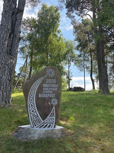 The image shows a stone monument in Pastavy, Belarus, that commemorates the year 2026 as the "Cultural Capital of Belarus." The monument is made of dark granite and features intricate carvings in white. At the top, a star-like motif is present, with a crescent-shaped design below it containing geometric patterns and vertical lines. The text "2026 ПАСТАВЫ КУЛЬТУРНАЯ СТАЛІЦА БЕЛАРУСІ" is inscribed on the stone. The monument is placed on a larger, lighter-colored stone base.
The monument is situated outdoors in a grassy area with several trees. To the left, a large tree with rough, textured bark dominates the foreground. In the background, more trees are visible, with a blue sky and scattered clouds overhead. The overall environment suggests a park or a natural setting within the city of Pastavy. There are no people or animals visible in the image.