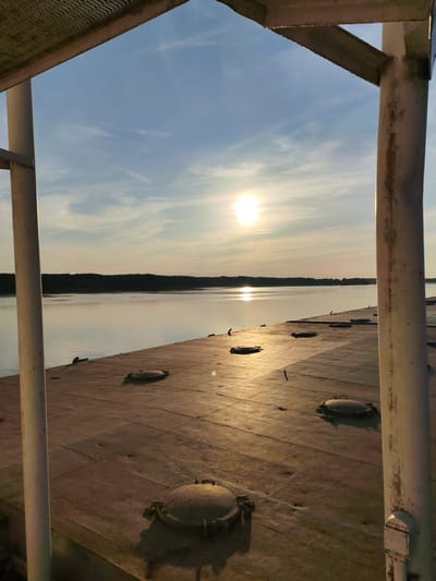 The image depicts a view from the deck of a barge or ship at sunset. The sun, a bright disc, is visible in the sky, casting a golden glow across the water and the deck. The water stretches out to a tree-lined horizon, and the sky is a pale blue with wispy clouds. The deck of the barge is made of large, flat panels and has several circular hatches, some of which are closed and some are slightly ajar. There are also some dark, curved objects that appear to be cleats or mooring points. The scene is framed by white metal poles and a slatted roof structure, suggesting the viewpoint is from under a covered area on the barge. The location is identified as the city of Shchetinino, Russia. There are no people visible in the image.