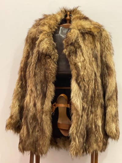 In an interior setting in Paiporta, Spain, a medium-length, shaggy fur coat, primarily in shades of light and dark brown, is displayed. The coat hangs open, revealing a dark brown, possibly leather, garment worn by a wooden mannequin or suspended from a hanger. Below this garment and partially obscured by the fur, a single tan-colored strappy wedge sandal with an ankle buckle is visible. The background is a plain, light-colored wall.