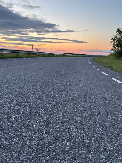 This is a low-angle, eye-level, outdoor, full shot of a paved road at sunset. The road curves to the right and dominates the foreground, with its rough, dark grey surface filling most of the frame. White dashed lines are visible on the right side of the road, indicating a lane or edge. To the left of the road, a metal guardrail runs along a grassy verge. Beyond the guardrail, the sky displays a gradient of colors, transitioning from pale blue at the top to hues of orange, pink, and yellow near the horizon, indicative of a sunset. Scattered clouds are present in the sky, some dark grey and others appearing lighter. In the distance, on the left side of the road, a signpost is visible. On the right side, a tree with green foliage stands beside the road. The overall environment appears to be rural or a roadside setting, likely within the city of Paldiski, Estonia, given the context provided. There are no people or specific actions depicted.