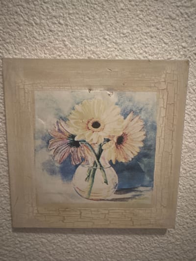 The image shows a framed painting of a bouquet of gerbera daisies in a clear glass vase. The flowers are predominantly yellow and pink. The background of the painting is a soft blue. The frame is a light, distressed wood with visible cracking. The painting is mounted on a textured, off-white wall. The scene is indoors, likely a home or establishment. There is no indication of the specific location within Santa Pola, Spain.