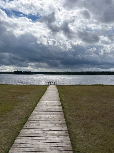 A wooden boardwalk extends from the foreground into a calm body of water, leading towards a distant tree line. The boardwalk is constructed from weathered wooden planks laid horizontally and appears to be situated on a grassy embankment. The body of water reflects the sky, which is dominated by dramatic, dark clouds interspersed with patches of brighter sky. The far bank of the water is lined with dense trees. The scene appears to be a natural landscape, possibly a lake or large river, with no visible buildings or urban structures.