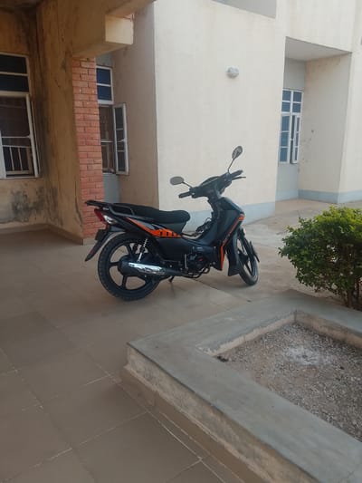 Outside a cream-colored building with multiple windows is a black and orange Lifan motorcycle parked on brown tiled ground. A small bush grows in a concrete planter next to the motorcycle. The building has both brick and plaster elements, and the lighting is natural daylight. The specific location within the city of Panhauya, Nigeria, cannot be determined. No people are visible.