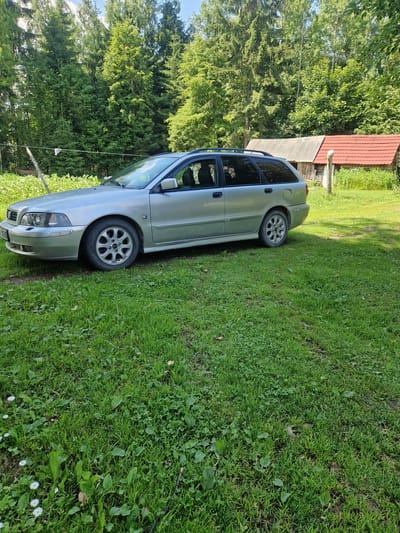 A silver station wagon is parked on a grassy area in front of two outbuildings with red and gray roofs. The outbuildings are set against a backdrop of dense green trees, likely a forest. The station wagon is facing left and its front bumper, headlights, and a portion of the driver's side are visible. The grass is well-maintained and appears to be a lawn, with some scattered daisies in the foreground. The sky is partly cloudy. The scene suggests a rural or suburban setting, possibly a residential property. This location is in the municipality of Rikava, Latvia.