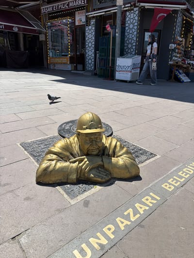 The image displays a vibrant street scene in Eskisehir, Turkey, illuminated by bright daylight, casting distinct shadows. The central subject is a golden statue of an elderly man, possibly depicting a traditional craftsman or vendor, positioned low to the ground on a black-patterned base. He appears to be kneeling or sitting, with his hands resting on his knees.
The statue is situated on a paved sidewalk, adjacent to a strip of golden lettering embedded in the ground that reads "UNPAZARI BELEDİ," likely part of "Odunpazarı Belediyesi," the local municipality. To the left of the statue, a pigeon pecks at the ground.
Along the street, several storefronts line the buildings. A red Turkish flag, bearing a white crescent and star, is prominently displayed above one shop entrance. Near this shop, a person stands by a white cooler, seemingly engrossed in their phone or another handheld device. The cooler is labeled "KALABAK DOĞAL KAYNAK SUYU" (Kalabak Natural Spring Water) and "External Spring Water." The building features a decorative white lattice pattern on its facade.
Further along, another shop, "BAŞAK ET PİLİÇ MARKET" (Başak Meat and Poultry Market), is visible with brightly lit signage. Other signs detail available products and services: "MUHTARIN YERI" (Headman's Place/Office) and a list of dairy items including "YOĞURT-KAYMAK TOST PEYNİRİ BEYAZ PEYNİR-KREMA TEREYAĞ" (yogurt, clotted cream, toast cheese, white cheese, cream, butter), explicitly stating "SÜT VE SÜT ÜRÜNLERİ BURADA SATILMAKTADIR" (milk and dairy products are sold here). The scene captures a slice of daily life in a lively urban setting.