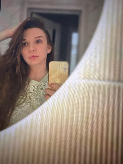 A young woman with long, brown hair is reflected in a mirror, taking a selfie with a gold-colored smartphone. She is wearing a light green top with a lace-like pattern. The mirror's edge is visible on the right, with a textured, ribbed surface. The background of the room is out of focus, showing a doorway and a wall. The image appears to be taken in a bathroom. The location is Засечное, Russia.