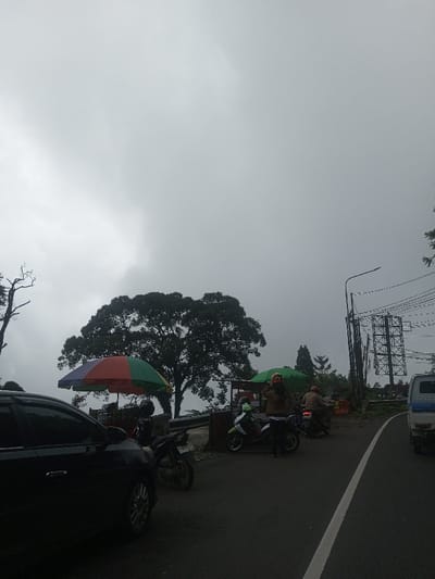 An overcast sky looms over a roadside scene. A black car is visible on the left, alongside parked motorcycles. A large tree stands nearby, partially obscured by a colorful umbrella. Further down the road, people gather around small food stalls shaded by green umbrellas. More motorcycles are parked along the road. Power lines and a metal structure are on the right side of the road, and a white van is visible in the distance.
