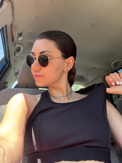 The image shows a close-up, angled portrait of a person (likely female) sitting in the front passenger seat of a car. The person has dark hair pulled back, wears dark round sunglasses, and has multiple ear piercings including a hoop, a stud, and a bar, along with a small hoop nose piercing. A blue and silver beaded necklace is visible around their neck. They are wearing a black sports bra or tank top. Their fingernails are painted white, with small black dots on the ring fingers.
Bright sunlight strongly illuminates the person's face, highlighting freckles, and their left arm, which is resting on a car seat. The car's interior, featuring a light-colored, somewhat worn headliner, a grab handle, and a sun visor, is visible. Through the car window, blurred outdoor scenery with green foliage and a hint of a red triangular road sign can be seen. The overall impression is a sunny daytime setting inside a vehicle. No discernible text is visible within the image. The location context provided is Baylovo, Bulgaria.