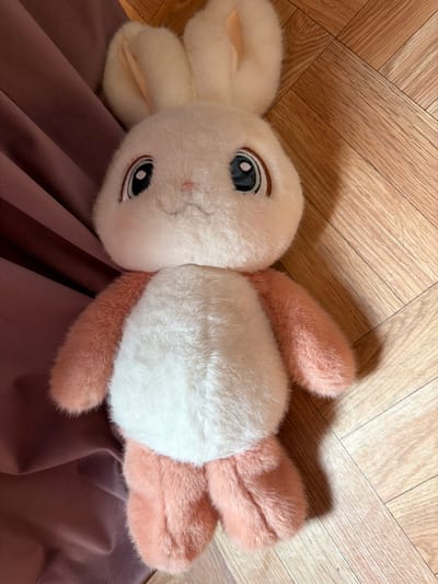 A plush bunny toy is lying on its back on a light brown wooden floor. The bunny has a white face, chest, and belly, with pink ears, arms, and legs. Its eyes are large and dark with brown irises. To the left of the bunny is a draped piece of dusty rose fabric. The scene is indoors, and the lighting suggests it is daytime. The image was taken in Verkhnevilyuysk, Russia.