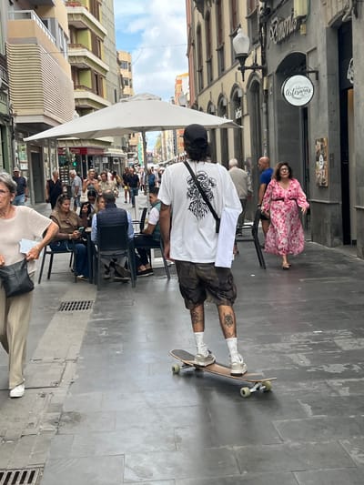 The image captures a street scene in Las Palmas De Gran Canaria, Spain. The location appears to be a pedestrian area lined with buildings. Several people are present, some seated at outdoor tables under a large white awning, likely part of a cafe or restaurant. One person is standing on a skateboard, and another is seated nearby. One woman in a pink floral dress is seated at a table, while other people are walking by. The street surface is tiled with what appear to be light-grey slabs. The sky is partly visible. There are streetlights and hanging signs along the buildings. The scene is taken at street level.