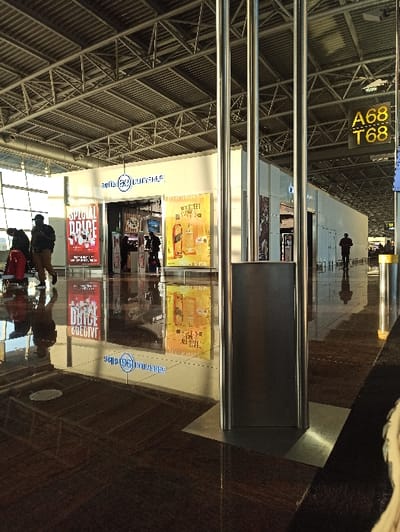 The image displays an interior view of an airport concourse in Steenokkerzeel, Belgium. A "belfio (36) DUTY FREE" shop with a white facade and a prominent blue circular logo is centrally located. To the left of the shop's entrance is a red poster advertising "SPECIAL PRICE," and to the right is a yellow poster featuring a bottle of "WALKER" whiskey with "MAKE SWEET STORIES" text. The floor is a dark, highly polished, reflective surface, mirroring overhead structures, signage, and figures. The ceiling features exposed metal trusses and ductwork, illuminated by overhead lighting and natural light entering from large windows on the left.

In the left foreground, a person wearing a dark jacket and cap walks right to left, pulling a red wheeled suitcase. Another person is partially visible behind them. Several indistinct figures are present inside or near the duty-free shop's entrance. In the far right background, a lone individual in a dark coat and hat walks away from the camera, their silhouette and clear reflection visible on the floor. Three polished metallic vertical poles stand in the right foreground. A yellow sign displaying "A68 T68" is mounted high on the wall to the right of the duty-free shop.