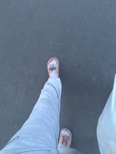 This is a first-person perspective image taken outdoors, likely on a paved surface such as asphalt, in Гожули, Ukraine. The focus is on the lower legs and feet of the individual. The person is wearing light grey sweatpants and light pink slides with a cartoonish graphic on the right slide. The left leg is bent at the knee, with the foot slightly forward. To the right of the frame, a portion of a white, opaque plastic bag is visible. The ground appears textured with small specks, consistent with asphalt. The lighting suggests it is daytime, possibly late afternoon due to the shadows.