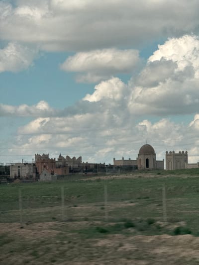 The image presents a panoramic landscape view, likely captured from a moving perspective, showcasing a settlement under a vast sky in Samsy, Kazakhstan.
The upper portion of the frame is dominated by an expansive blue sky interspersed with numerous large, fluffy cumulus clouds, varying in shades from bright white to soft gray. These clouds create a dynamic ceiling, with occasional clear patches of blue sky visible between them.
Stretching horizontally across the middle ground is a line of distinctive buildings. These structures exhibit an architectural style featuring light-colored facades, intricate detailing, and varied rooflines, reminiscent of Central Asian or Middle Eastern design. A prominent building features a large, beige-colored dome and what appears to be a smaller minaret or tower on its roof. Other buildings along the line display crenelated tops and multi-story constructions, suggesting a well-established town or complex.
The lower part of the image consists of a broad expanse of land. On the left, it appears as a green, grassy field, while the right side transitions into a drier, more rugged terrain, possibly an uncultivated area or a roadside. Faint horizontal lines are subtly visible across the grassy foreground, potentially indicating distant utility lines or field boundaries.
The scene is illuminated by daylight, suggesting a late morning or afternoon setting under partly cloudy conditions. No specific activities or individuals are observable within the frame, and there is no visible text. The image conveys a sense of open space and distant architectural presence within the Kazakh landscape.
