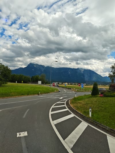 This media file captures a scenic view of a road curving through a grassy landscape with a majestic mountain range in the background. The scene appears to be set in Walserfeld, Austria.
The foreground features a paved road with clear white lane markings, indicating a junction or an approach to one. The road curves to the right, with a grassy embankment and a landscaped area to its side. Several white bollards line the edge of the road, and there are also some small white rectangular markers on the road surface.
In the middle ground, a lush green field stretches out, bordered by trees. Beyond the field, a line of buildings can be seen, suggesting a small town or village. Traffic signs are visible along the road, including a red and white circular sign indicating "no entry" and a blue sign with white text, likely providing directional information.
The background is dominated by a large, dark mountain range, its peaks and slopes shrouded in a mix of sunlight and shadow. The sky above is a dynamic canvas of dramatic clouds, with patches of blue sky peeking through. Some of the clouds are heavy and dark, suggesting potential rain, while others are bright and white, illuminated by the sun.
A solitary street lamp stands on the right side of the road, its light directed downwards. There are no people visible in the image. The overall impression is one of natural beauty and open space, with the road serving as a pathway through the landscape. The weather appears to be changeable, with a mix of sunshine and heavy cloud cover, typical of a summer day in a mountainous region. The time of day is likely afternoon, given the lighting conditions. The location cues, such as the road layout and the specific appearance of the mountains, are consistent with a European setting, specifically Austria as indicated by the location context.