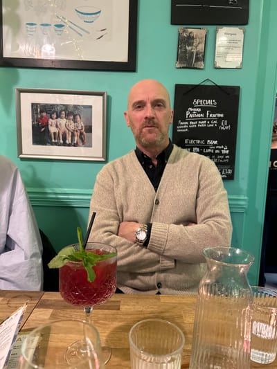 The image shows the interior of a restaurant or bar, possibly in Bath, United Kingdom, based on the presence of signs and the overall atmosphere. A man with a shaved head, wearing a beige cardigan over a dark shirt and a watch, is seated at a table with his arms crossed. A red cocktail in a patterned glass, garnished with mint and a black straw, sits on the table next to empty glassware, including a carafe. The table is wooden.
Behind the man is a light turquoise wall adorned with framed artwork and blackboards. The blackboards display the word "SPECIALS" and menu items, including a Mango Passion Fruitini and Electric Bear IPA. The wall also displays allergy information and other framed images. The overall environment suggests a casual dining or drinking establishment.