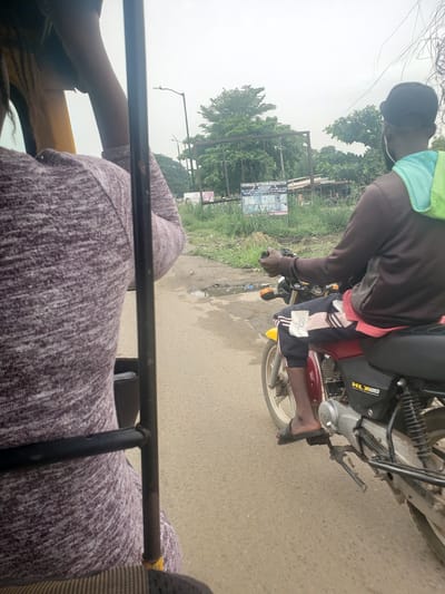 This image captures a roadside scene in Ida, Nigeria, from the perspective of a passenger in an open-sided vehicle, possibly a Keke NAPEP. The lighting is muted, indicating an overcast day.

The central subject is an individual riding a black and red motorcycle, with "HLX 3" visible on the engine casing. The rider is wearing a dark long-sleeved top, dark pants with white stripes and a white logo on the left thigh, and a light green neck gaiter or bandana. Dark earbuds or headphones are visible. The rider's dark-skinned bare feet are visible, one resting on the motorcycle's footrest, adorned with dark sandals.

In the foreground, a black metal bar, part of the photographer's vehicle, spans horizontally across the lower section. The photographer's arm and shoulder, wearing a purple and white patterned long-sleeved garment, are partially visible.

The setting includes a paved road with some dirt and debris. Along the left side of the road, there's a grassy area with various trees and bushes. A large billboard stands prominently here, displaying the text "SHANDARA INTERIOR DECORATION." Smaller text below specifies their services: "WE ARE EXPERTS IN ALL KINDS OF INTERIOR, EXTERIOR, PLUMBING, PAINTING, POP, AND TYLING WORK. WE DEAL IN ALL KINDS OF PIPES." Images related to interior design are also visible on the sign. Overhead wires are discernible against the bright, featureless sky. The scene depicts typical daily transit in a West African locale.