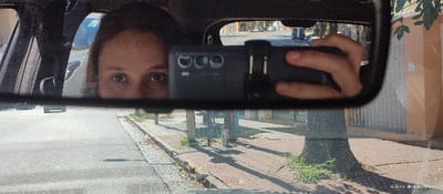 The image displays a scene from inside a vehicle, centered on the reflection within its rearview mirror. The mirror shows the upper portion of a female individual's face, specifically her eyes and forehead, positioned to the left. A dark-colored smartphone, held horizontally by a human hand, is reflected to the right, partially obscuring the lower face of the individual. The phone features at least three distinct rear camera lenses.
Outside the vehicle, through the windshield, an urban street scene in Quilmes, Argentina, is visible. The foreground shows an asphalt road and a concrete sidewalk on the right, sparsely covered with dry leaves and patches of grass. A tree with a textured trunk stands on the sidewalk. In the midground, several buildings line the street, including a light orange/beige structure with a white security grille on its window. The presence of distinct shadows on the sidewalk indicates bright daylight. A distant, light-colored vehicle is partially visible on the road outside the mirror's frame. The overall composition suggests a self-portrait taken by the individual using the smartphone and the car's rearview mirror.