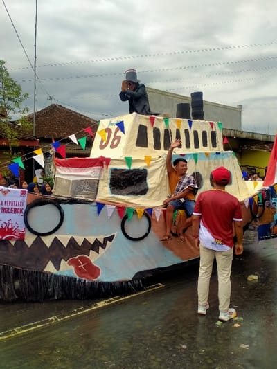 The image captures a daytime outdoor scene in Mojogedang, Indonesia, showcasing a large, elaborate parade float positioned on a wet paved road beneath an overcast sky. The float's lower section is painted to resemble a creature's head, featuring dark jagged teeth, two black circular designs, and a prominent red tongue. The upper structure is a light-colored cabin-like section with painted dark rectangular windows and the brown number '06' on its side. Multicolor triangular pennants adorn both parts of the float.
On the highest point of the cabin, a male figure wearing a dark suit and a grey top hat is seen bowing with his hands pressed together. A young male, dressed in an orange shirt and blue shorts, is seated on the cabin's side, smiling and waving his right hand. A white banner with red text, partially visible as 'DESA BERKREASI ANGLEBAK,' is attached to the left front of the float.
In the foreground, a male individual in a red and white short-sleeved shirt, light-colored trousers, and sneakers stands with his back to the viewer, observing the float. In the background to the left, a group of people, including several women wearing hijabs, are partially visible, observing the scene from behind a smaller banner. Traditional tiled-roof buildings and utility lines are visible against the cloudy horizon. The wet pavement reflects the overhead light, indicating recent rainfall or standing water.