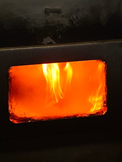The scene, located in Opaka, Bulgaria, depicts a close-up of vigorous combustion contained within a dark, metallic appliance, likely a wood-burning stove or fireplace. A prominent horizontal rectangular window, centrally positioned, reveals bright orange and yellow flames actively burning. The flames undulate and fill the majority of the visible glass area. The window's perimeter displays a textured, possibly sooted or heat-affected appearance. The surrounding appliance surface is dark and exhibits minor surface irregularities above the window. No individuals or additional objects are present in the frame.